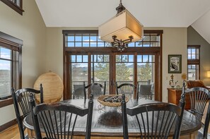 A dining area with a table, chairs, a hanging light fixture, and large windows with a view. - New! Cozy Craftsman Condo | Walk to Downtown Whitefish, Gas Fireplace & Mountain Views (Whitefish)