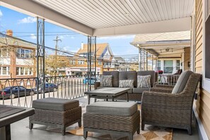 A covered outdoor seating area with wicker furniture, a table, and a view of the street and buildings. - Modern 6BR | Sleeps 14 | 1.5 Blks to Beach (Ventnor City)