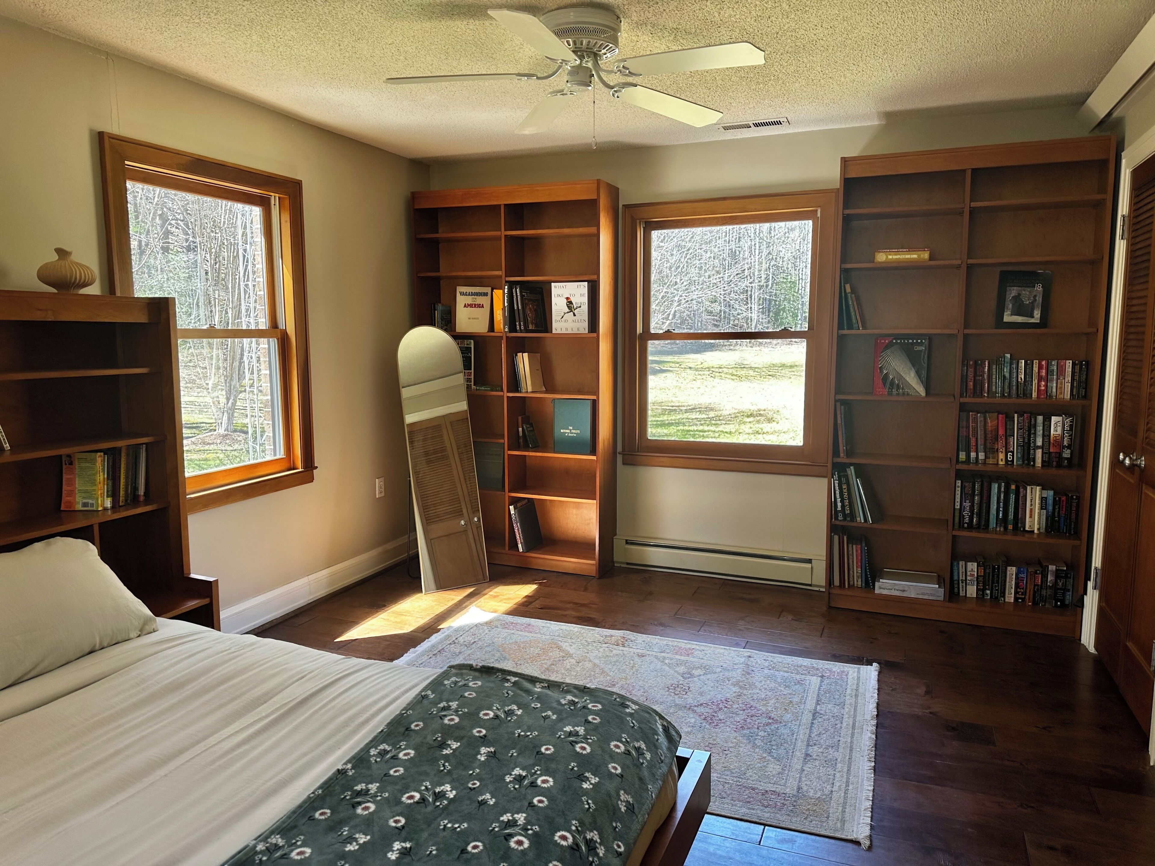 A bedroom with a bed, a standing mirror, a ceiling fan, and bookshelves.