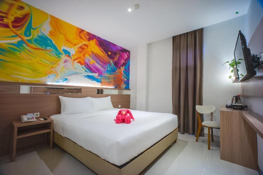 Habitación de hotel con una cama grande, una mesita de noche con teléfono, un escritorio con silla, un televisor montado en la pared y un cuadro abstracto de colores sobre la cama.