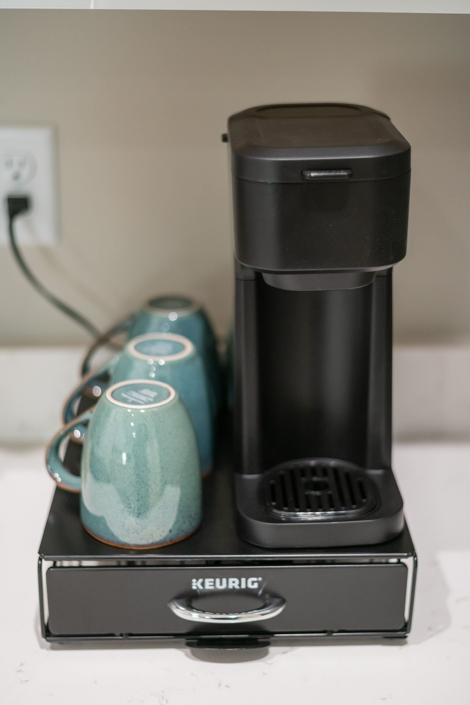 Una cafetera Keurig sobre una base negra, con tres tazas color turquesa a su lado.