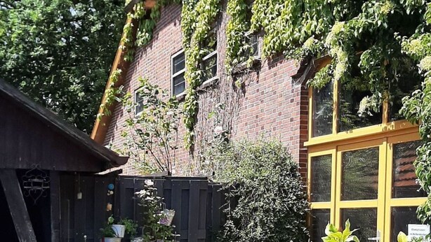 Ein Garten mit einem Backsteinhaus, einer Holzhütte und unterschiedlichen Pflanzen.