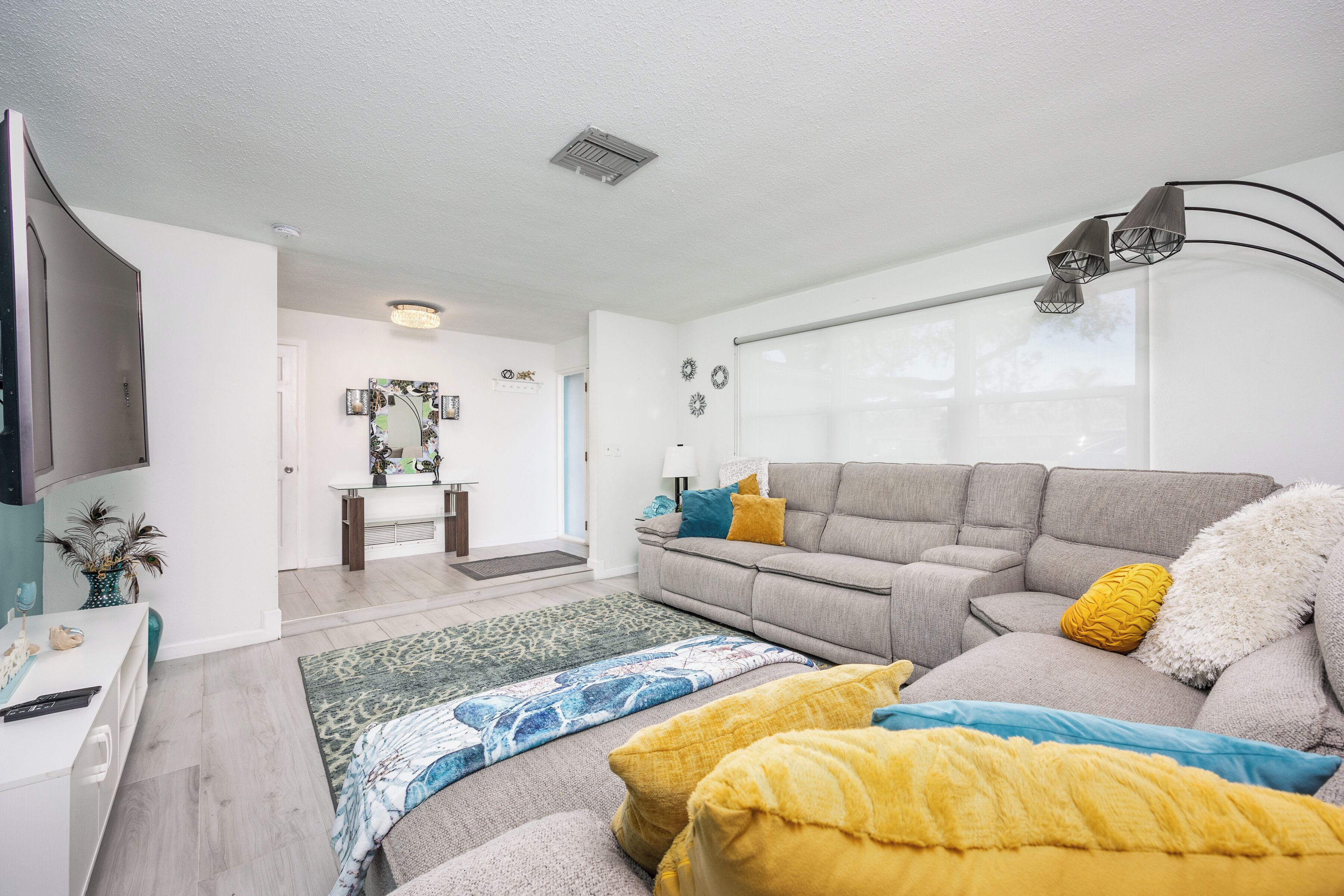 Ein modernes Wohnzimmer mit einem grauen Sofa, gelben und blauen Kissen, einem gemusterten Teppich und einem Flachbildfernseher.