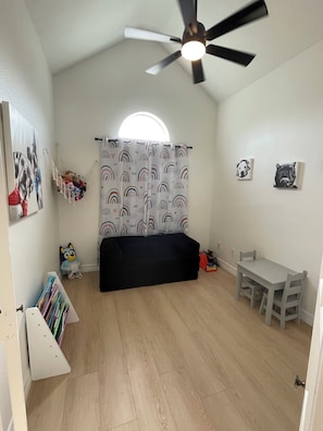 A room with a ceiling fan, a black sofa, a small table, a bookshelf, and a window with a curtain.