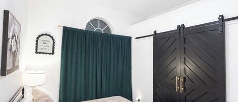 A bedroom with a bed, a dark sliding door, a green curtain, a wall clock, and a framed quote.