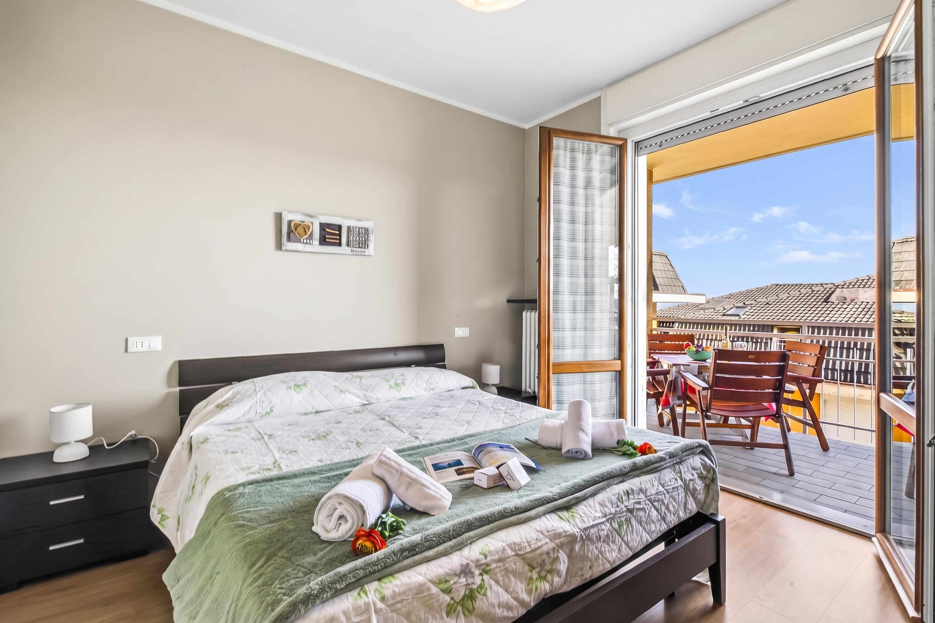 A hotel room with a bed, bedside table, lamp, and a view of the outside balcony.