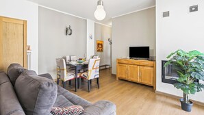 Ein Wohnzimmer mit grauem Sofa, Esstisch, Stühlen, einem Fernseher, Holzschränken und einem Blumentopf.