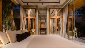 Una cama grande con colcha blanca y varios almohadones, una puerta de vidrio que da a un balcón y vistas al exterior.