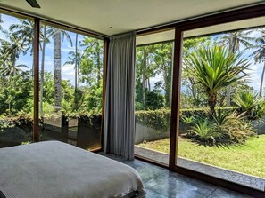 A bedroom with a large bed, a window with curtains, and a view of a tropical garden. - Beachside Villa at Balian Surf Break (Selemadeg Barat)
