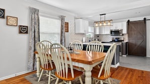 A dining area with a wooden table and white chairs, a kitchen with white cabinets and a dark wooden door, and a window with blinds. - Premier Louisville home with exclusive home gym! (Louisville)