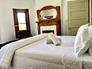 Ein ordentlich bezogenes Bett mit gesteppter Decke, ein aufgerolltes Handtuch und ein Kopfkissen. Im Raum befindet sich ein Holzstuhl, ein Spiegel und eine Tür.