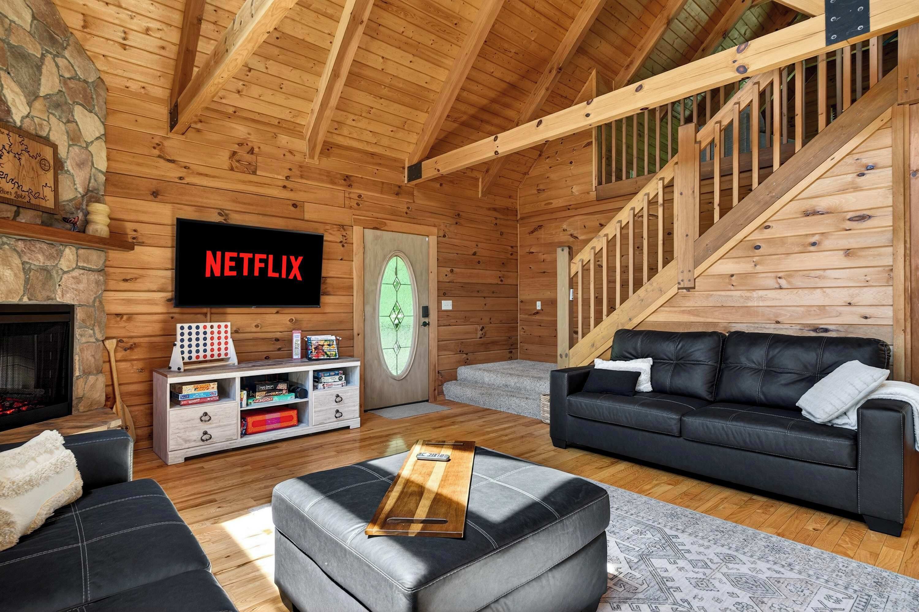 Un accogliente interno di una baita in legno, con un camino, un divano in pelle nera, un mobile TV in legno e una scala.