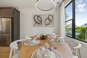 A modern dining area with a wooden table set for six, white chairs, and a view of greenery through a large window. - Iconic retreat w/ exclusive rooftop lounge & spa! (Nashville)