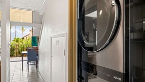 Un cuarto de lavandería con lavadora y secadora, una puerta que da a un balcón con vista a zonas verdes, y una silla.
