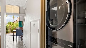 Un cuarto de lavandería con lavadora y secadora, una puerta que da a un balcón con vista a zonas verdes, y una silla.