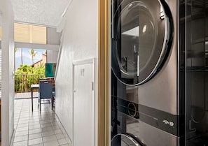 A laundry room with a washing machine and dryer, a door leading to a balcony with a view of greenery, and a chair. - Cozy 2-bedroom condo with AC in lovely Kihei (Kihei)