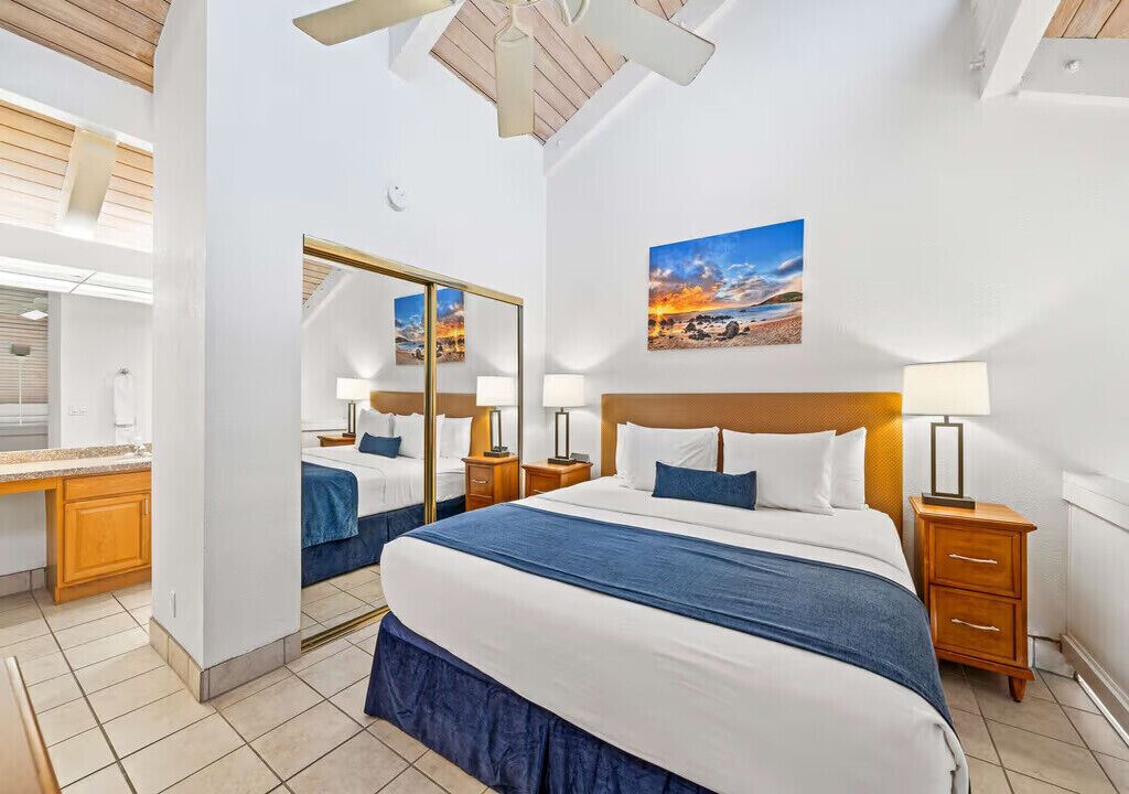 Une chambre d’hôtel avec un grand lit, un miroir, une table de chevet avec une lampe, un tableau accroché au mur et un ventilateur de plafond.
