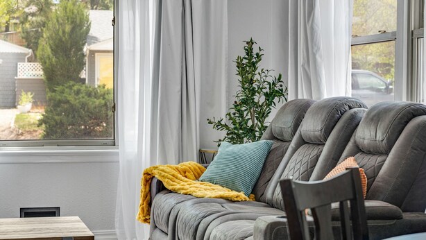 A cozy living room with a grey sofa, a small table, and a window with curtains. - Bright Beautiful 2BR Near Broadmoor with King Bed, Jetted Tub, Yard & Deck (Colorado Springs)
