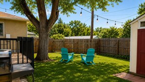 A backyard with a wooden fence, two turquoise Adirondack chairs, a barbecue grill, and string lights. - Bright Beautiful 2BR Near Broadmoor with King Bed, Jetted Tub, Yard & Deck (Colorado Springs)