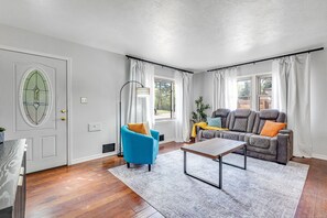 A living room with a blue armchair, a gray sofa, a wooden coffee table, and a large window with curtains. - Bright Beautiful 2BR Near Broadmoor with King Bed, Jetted Tub, Yard & Deck (Colorado Springs)