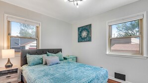 A bedroom with a bed, a nightstand, a lamp, a wall art piece, and windows. - Bright Beautiful 2BR Near Broadmoor with King Bed, Jetted Tub, Yard & Deck (Colorado Springs)