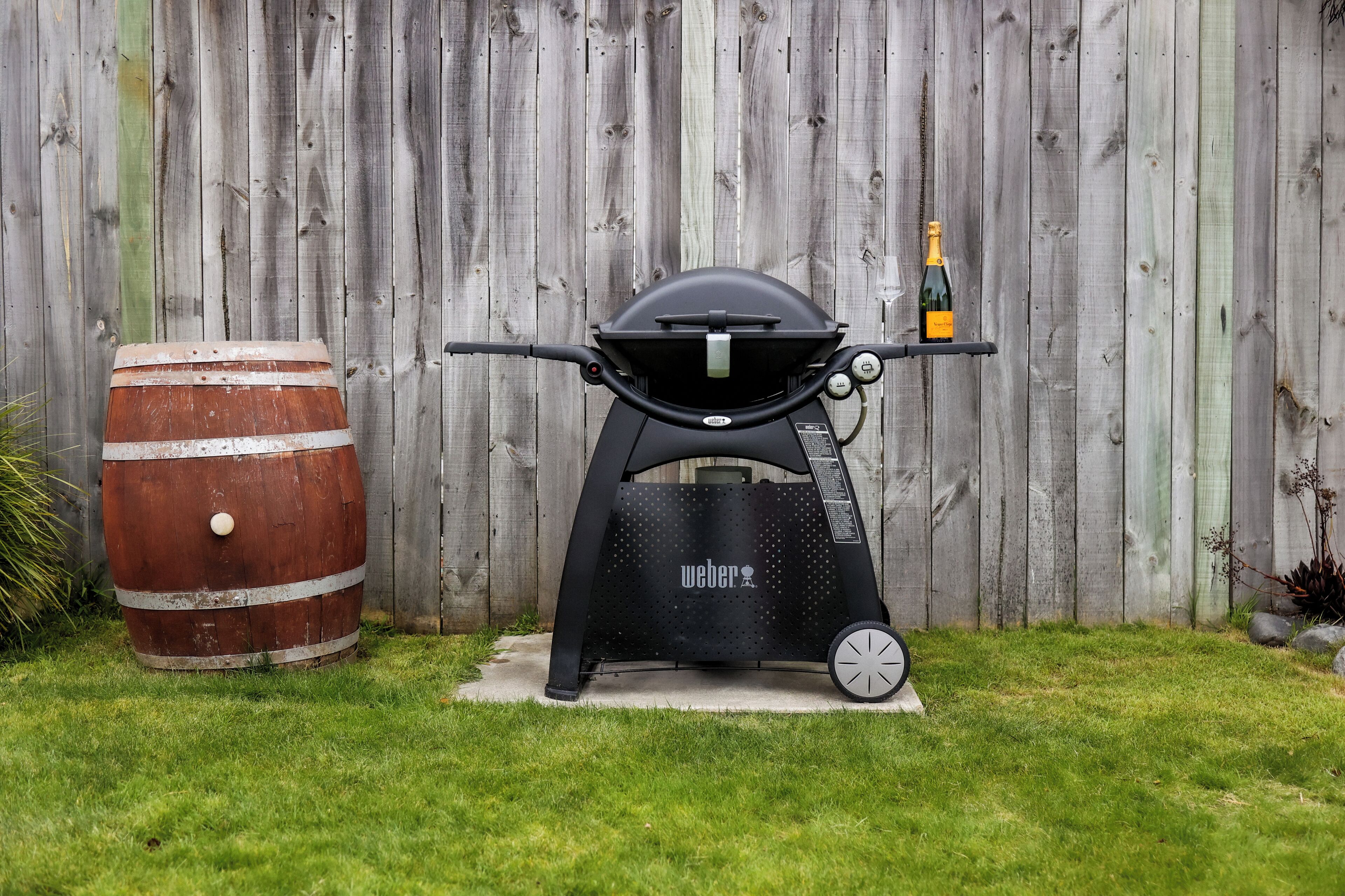 A Weber grill with a bottle of champagne, a wooden barrel, and a wooden fence in the background.