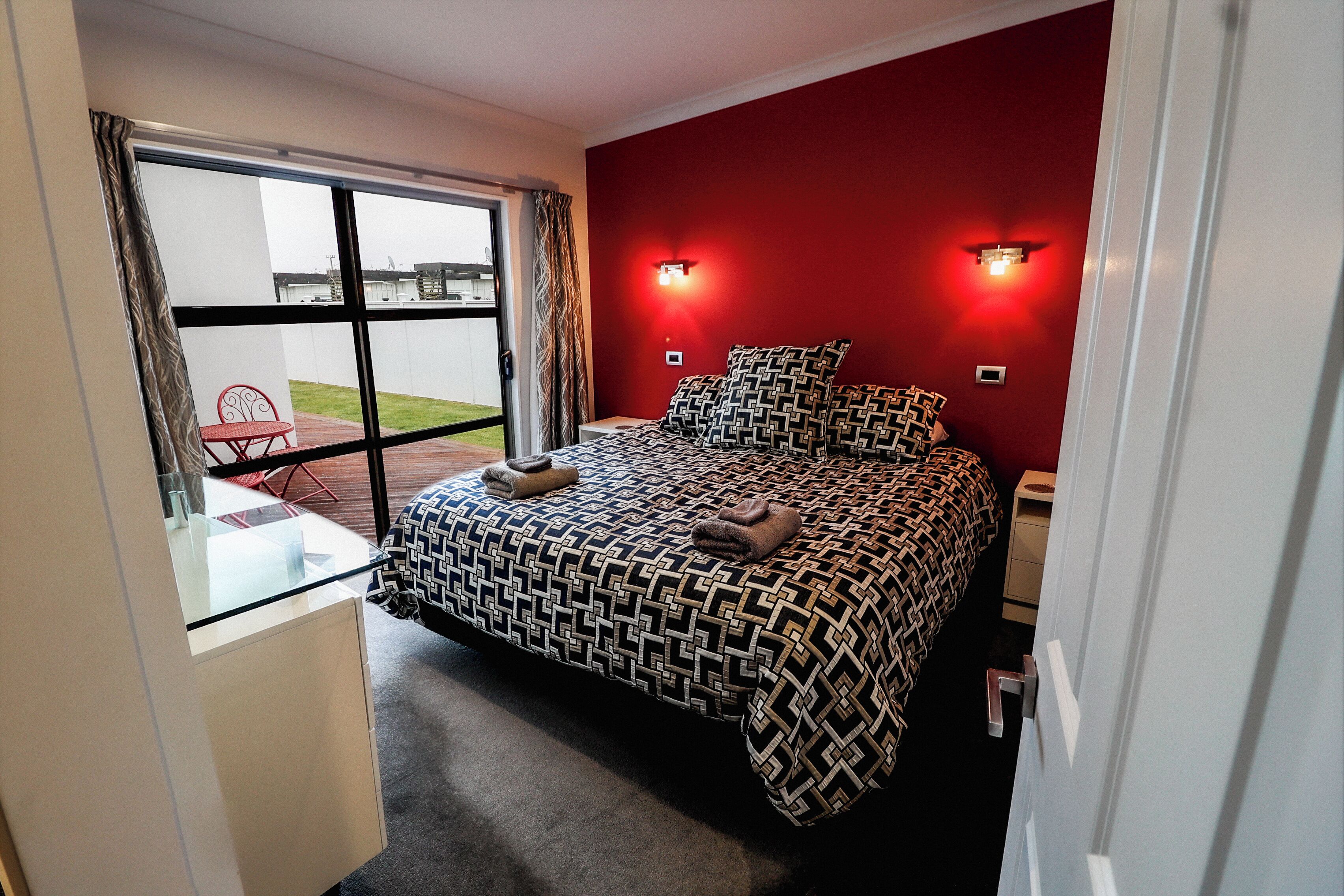 A bedroom with a bed, a desk, and a chair, a window with curtains, and a red wall with two wall lights.
