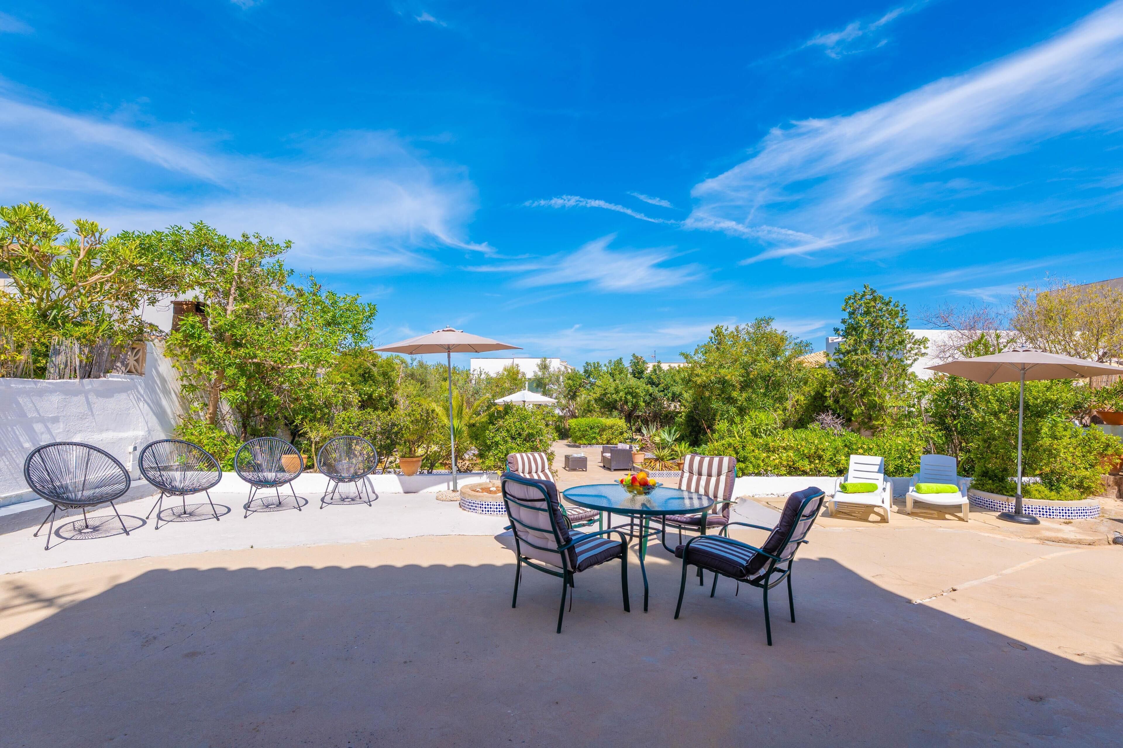 An outdoor patio with a round table and chairs, surrounded by greenery and umbrellas.
