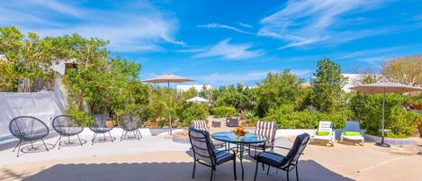 An outdoor patio with a round table and chairs, surrounded by greenery and umbrellas.