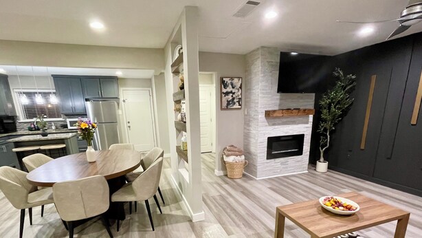 A modern kitchen with grey cabinets, a dining area with a wooden table and chairs, a fireplace, and a television mounted on the wall. - The Garland Getaway • Backyard Oasis • Family Friendly • Near Dallas (Garland)