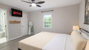 A bedroom with a bed, a ceiling fan, a TV displaying 'NETFLIX,' a window with blinds, and a door leading to a bathroom. - The Garland Getaway • Backyard Oasis • Family Friendly • Near Dallas (Garland)