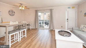 A living room with a ceiling fan, a dining table with stools, a sofa, and a small kitchen area with a sink. - Sinbad – Newly Renovated • Steps to the Beach • Midtown OC 2BR 1.5 BA (Ocean City)