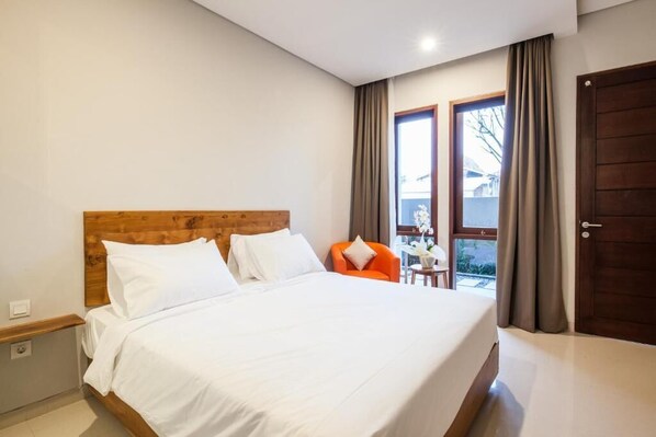 Une chambre à coucher avec une tête de lit en bois, un grand lit avec une literie blanche, un fauteuil orange, une petite table avec un vase, et une vue par une fenêtre avec des rideaux.