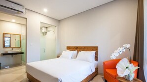 A modern bedroom with a wooden bed, a small orange sofa, and a bathroom with a glass shower enclosure. - Eclipse Kerobokan (Seminyak)