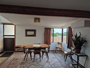 A dining area with a wooden table and chairs, a patterned rug, and a view through large windows.