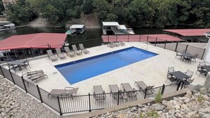 Una piscina con lettini e tavoli, circondata da una recinzione, con un fiume e una zona boschiva sullo sfondo.
