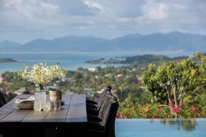Une table à manger ornée d’un vase de fleurs blanches, de verres et d’assiettes, donnant sur une piscine et offrant une vue panoramique sur les montagnes et la côte.