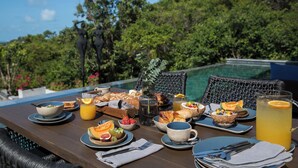 Une table dressée pour le petit-déjeuner, avec divers aliments et boissons, donnant sur une piscine et une végétation luxuriante.