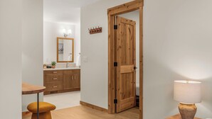A modern hotel room with a wooden door, a small table with a stool, a bedside lamp, and a mirror above the sink. - Alpine Forest Retreat Sauna + Hot Tub + EV (Canmore)