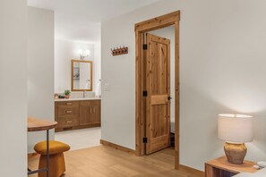 A modern hotel room with a wooden door, a small table with a stool, a bedside lamp, and a mirror above the sink. - Alpine Forest Retreat Sauna + Hot Tub + EV (Canmore)