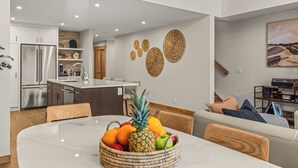 A modern kitchen with a stainless steel refrigerator, a dining table with a fruit bowl, and a living area with a sofa and a painting. - Alpine Forest Retreat Sauna + Hot Tub + EV (Canmore)