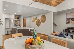 A modern kitchen with a stainless steel refrigerator, a dining table with a fruit bowl, and a living area with a sofa and a painting. - Alpine Forest Retreat Sauna + Hot Tub + EV (Canmore)