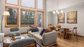 A modern living room with a sofa, chairs, a dining table, and large windows. - Alpine Forest Retreat Sauna + Hot Tub + EV (Canmore)