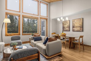 A modern living room with a sofa, chairs, a dining table, and large windows. - Alpine Forest Retreat Sauna + Hot Tub + EV (Canmore)