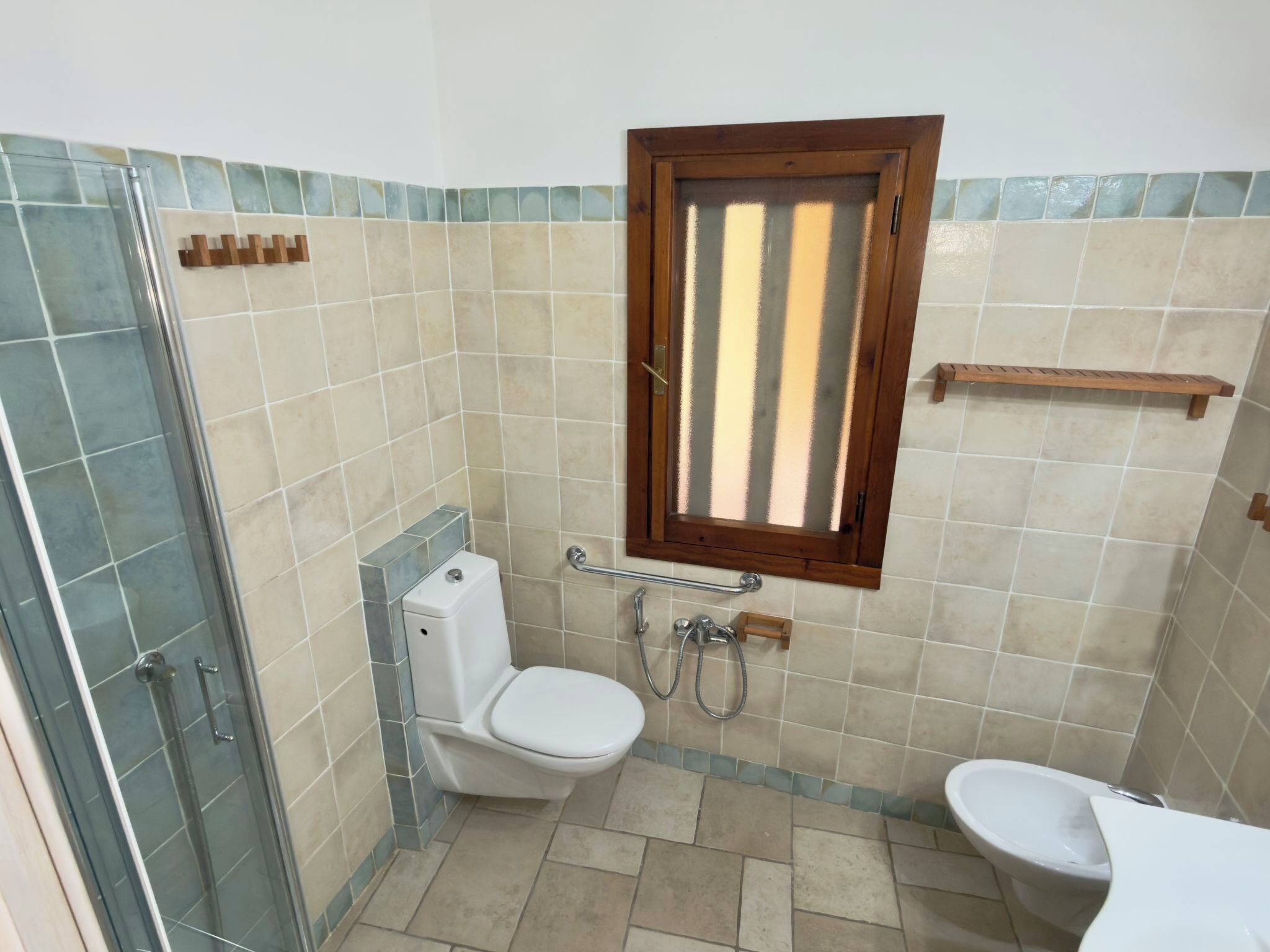 Ein Badezimmer mit Dusche, Toilette, Bidet und einem Fenster mit Jalousien.