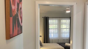 A bedroom with a bed, a window with curtains, a door leading to another room, and a painting on the wall.