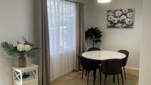 A dining area with a round table and chairs, a small white side table with a plant, a window with curtains, and a wall-mounted painting.
