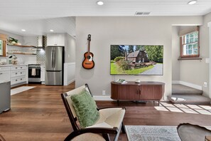 A modern kitchen with stainless steel appliances, a dining area with a wooden table, a comfortable seating area with a green cushion, and a large flat-screen TV mounted on the wall. - Cozy Conifer Cottage- A Newly Reimagined Woodland Retreat (Port Angeles)