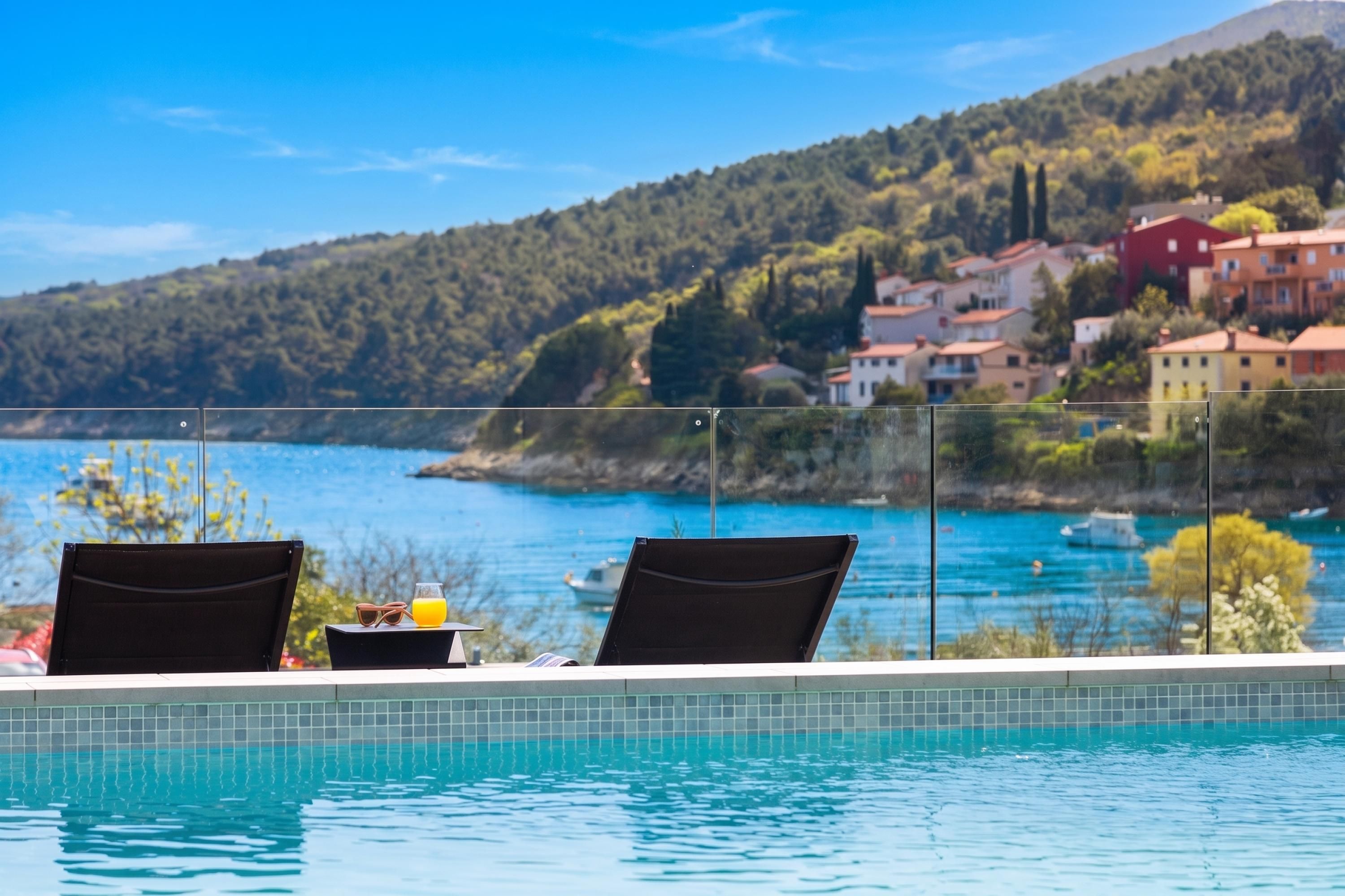 Vista junto a la piscina con dos sillas, una mesa con un vaso de zumo de naranja y una panorámica de la costa con edificios y colinas.