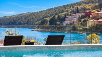 Vista junto a la piscina con dos sillas, una mesa con un vaso de zumo de naranja y una panorámica de la costa con edificios y colinas.